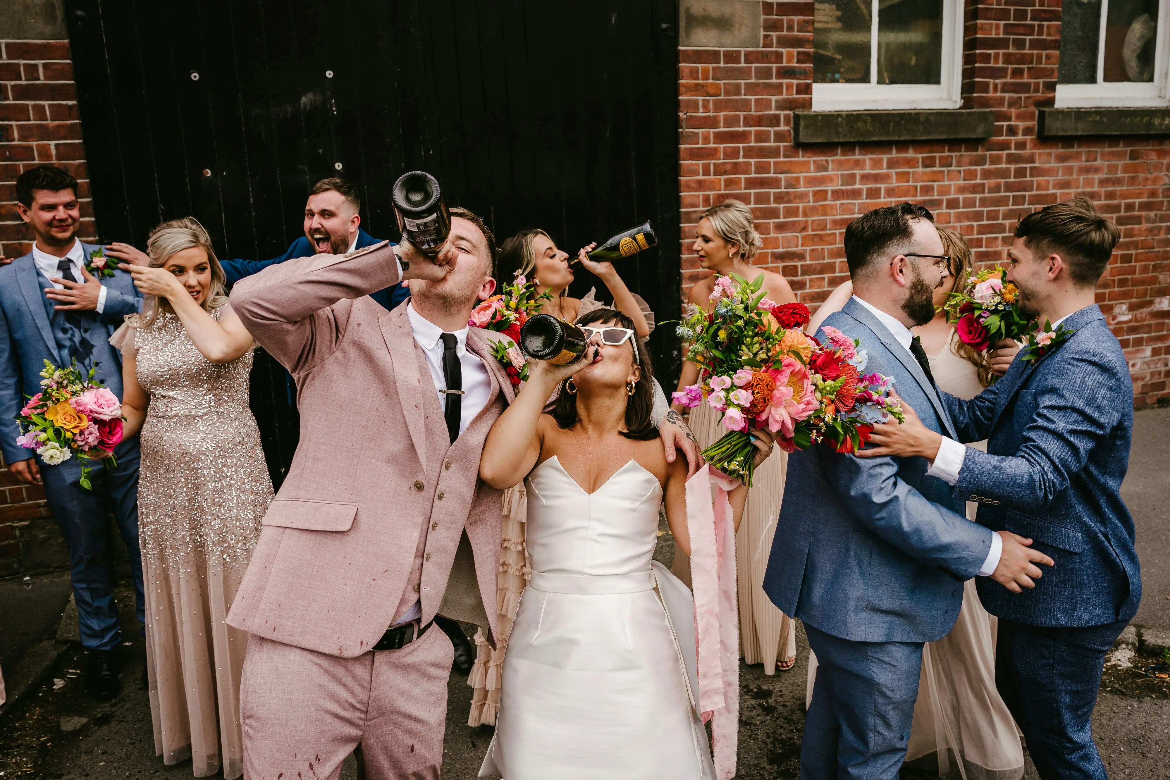 Bride, groom, and wedding party celebrating with champagne outside a brick venue.