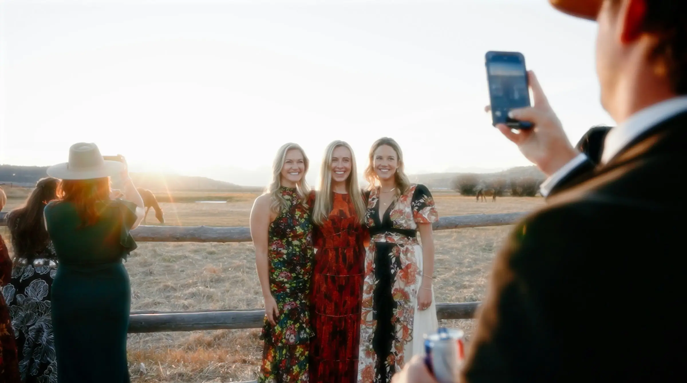 Wedding guest taking a smartphone photo of three women posing outdoors.