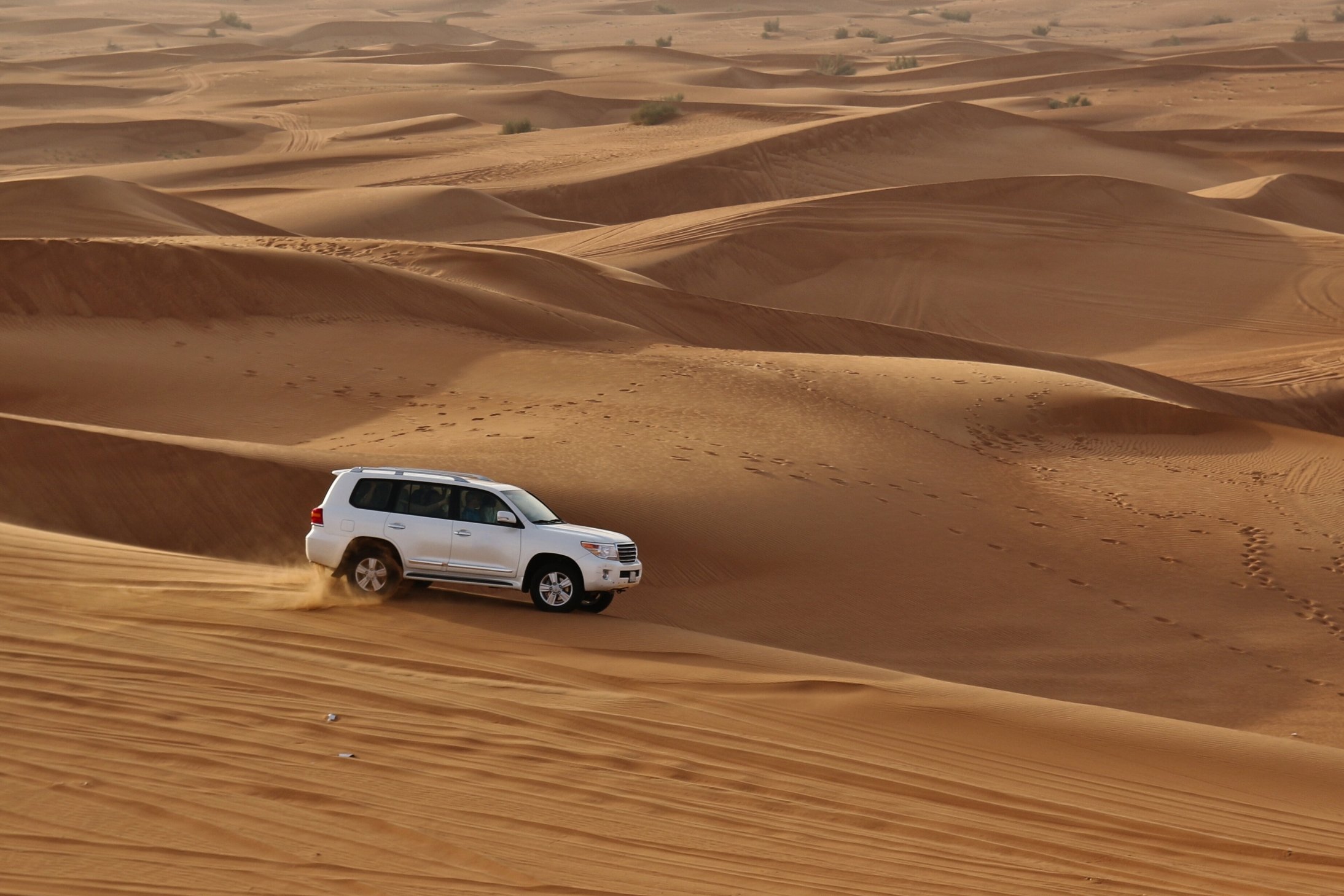 White SUV driving off-road on vast sand dunes in a desert landscape.