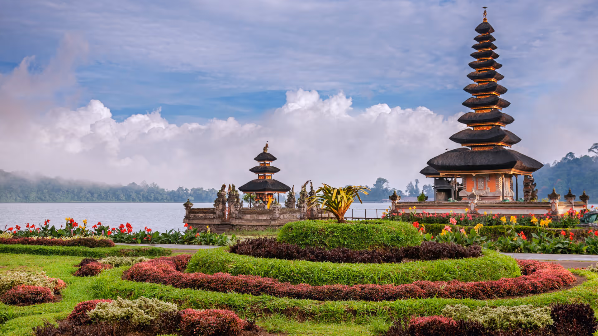 Traditional Balinese temple with tiered roofs by a lake, surrounded by manicured gardens and colorful flowers.