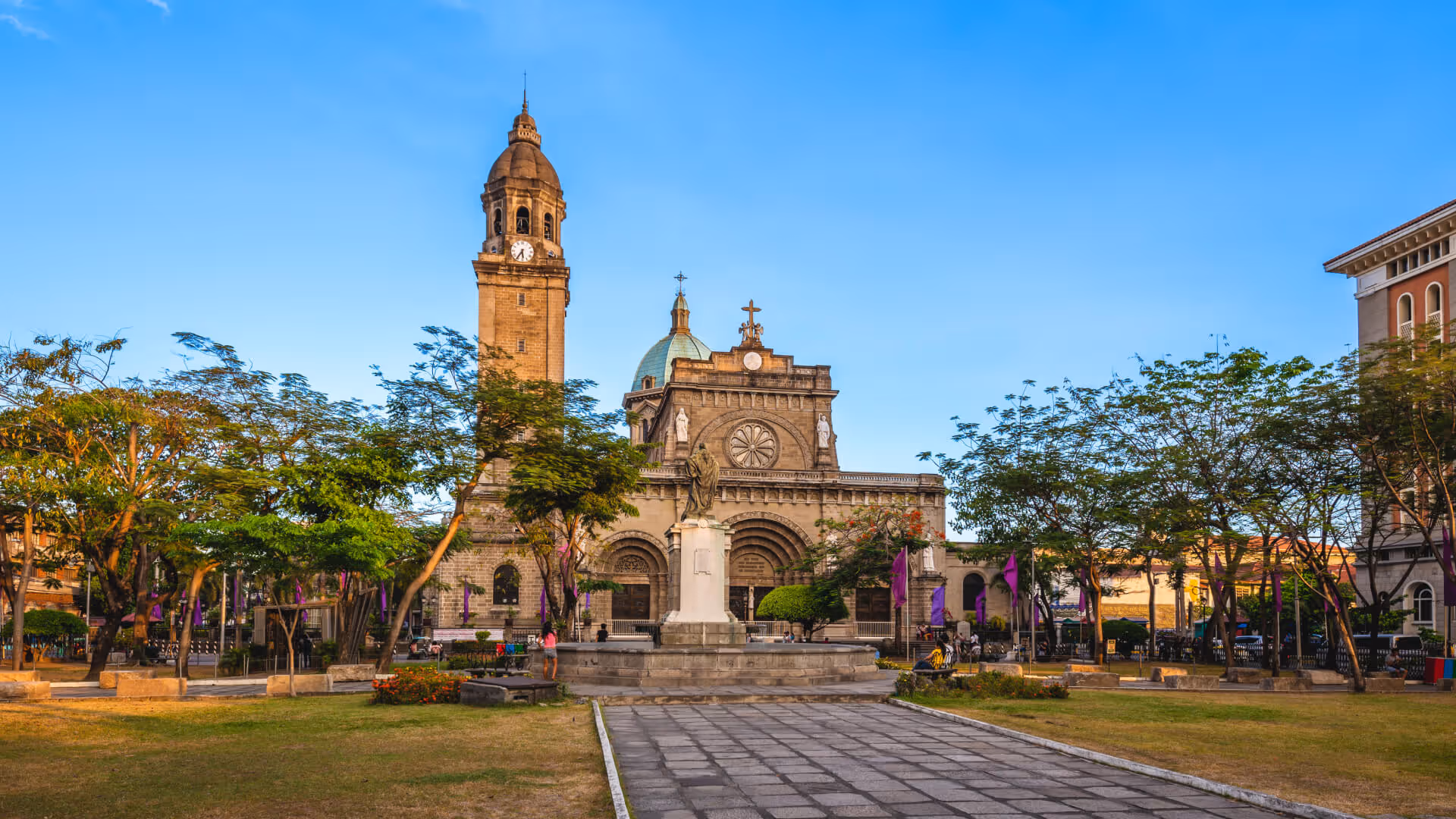 Manila Cathedral with clock tower and statue at the front, surrounded by trees and purple banners.