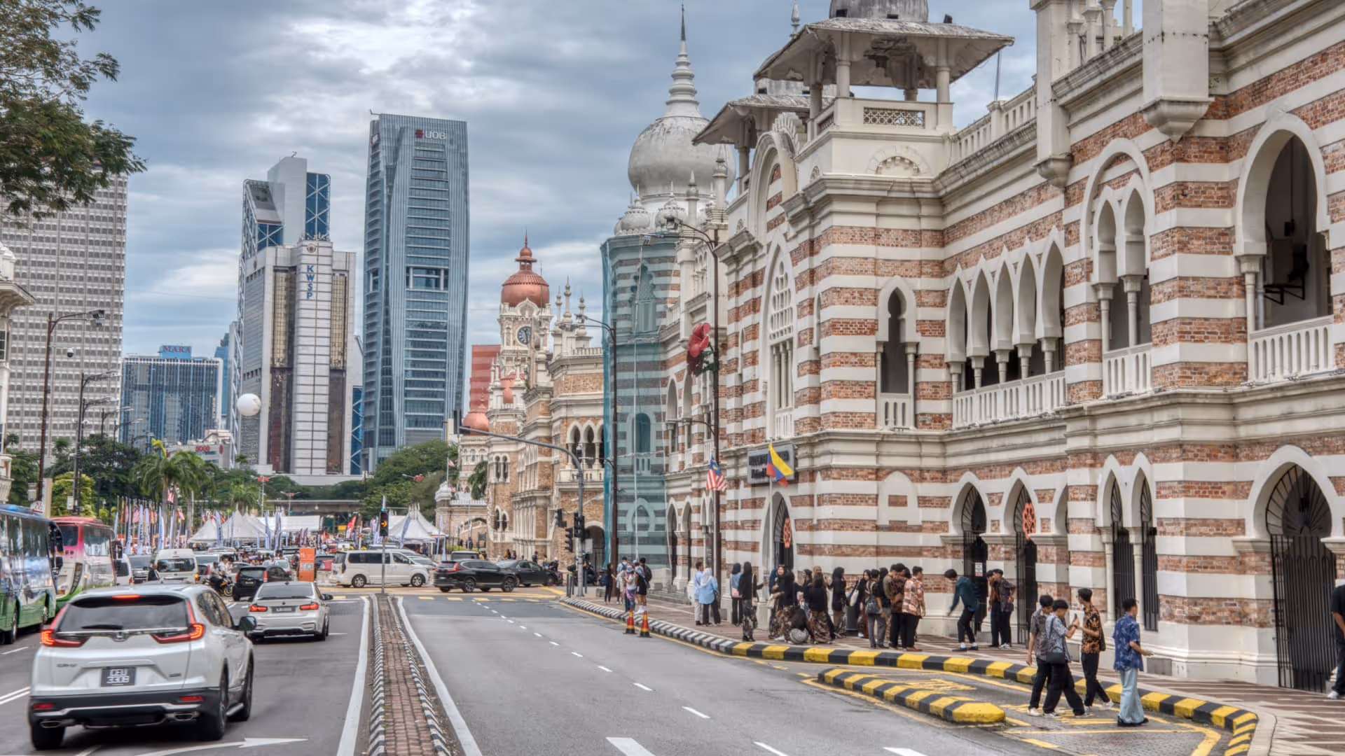 Street scene in Kuala Lumpur with traffic, pedestrians, and colonial-style buildings against modern skyscrapers.