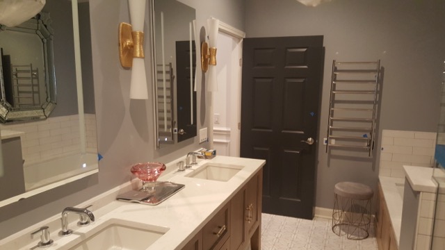 Elegant bathroom remodel by Chris Painting and Remodeling featuring gray walls, white subway tile, double vanity with marble countertop, gold sconces, large mirrors, and dark accent door