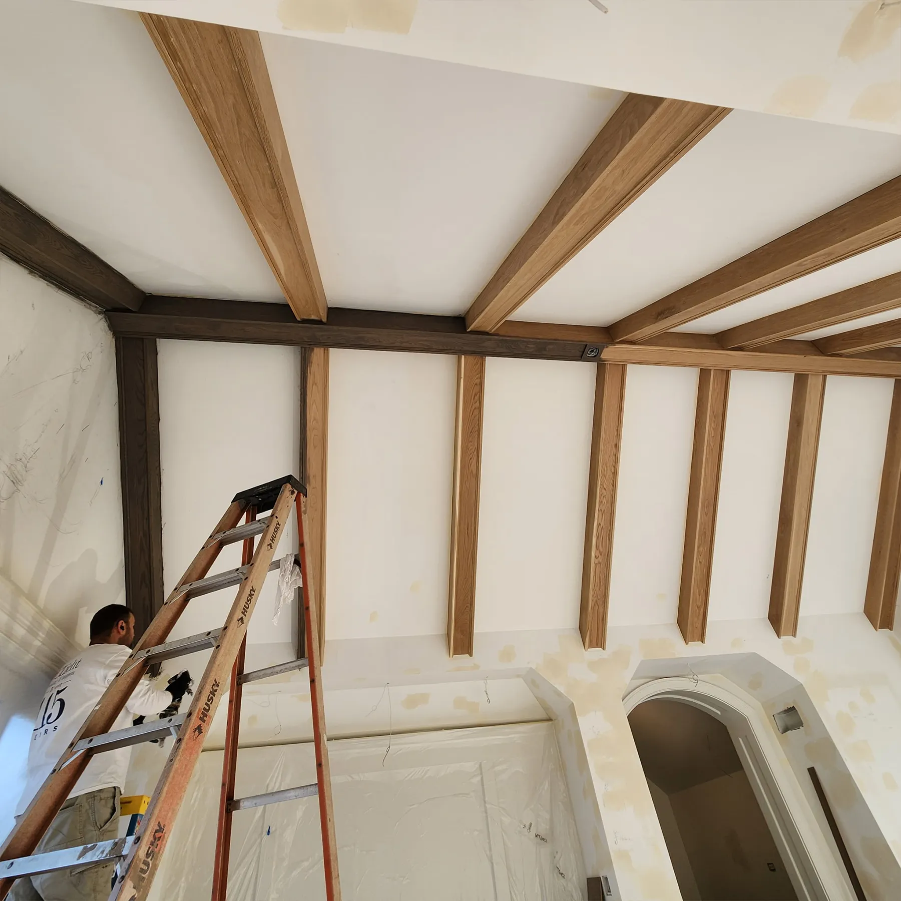 Construction view of vaulted ceiling with stained oak wooden beams and cross beams during installation by Chris Painting and Remodeling, worker on ladder applying finishing touches.