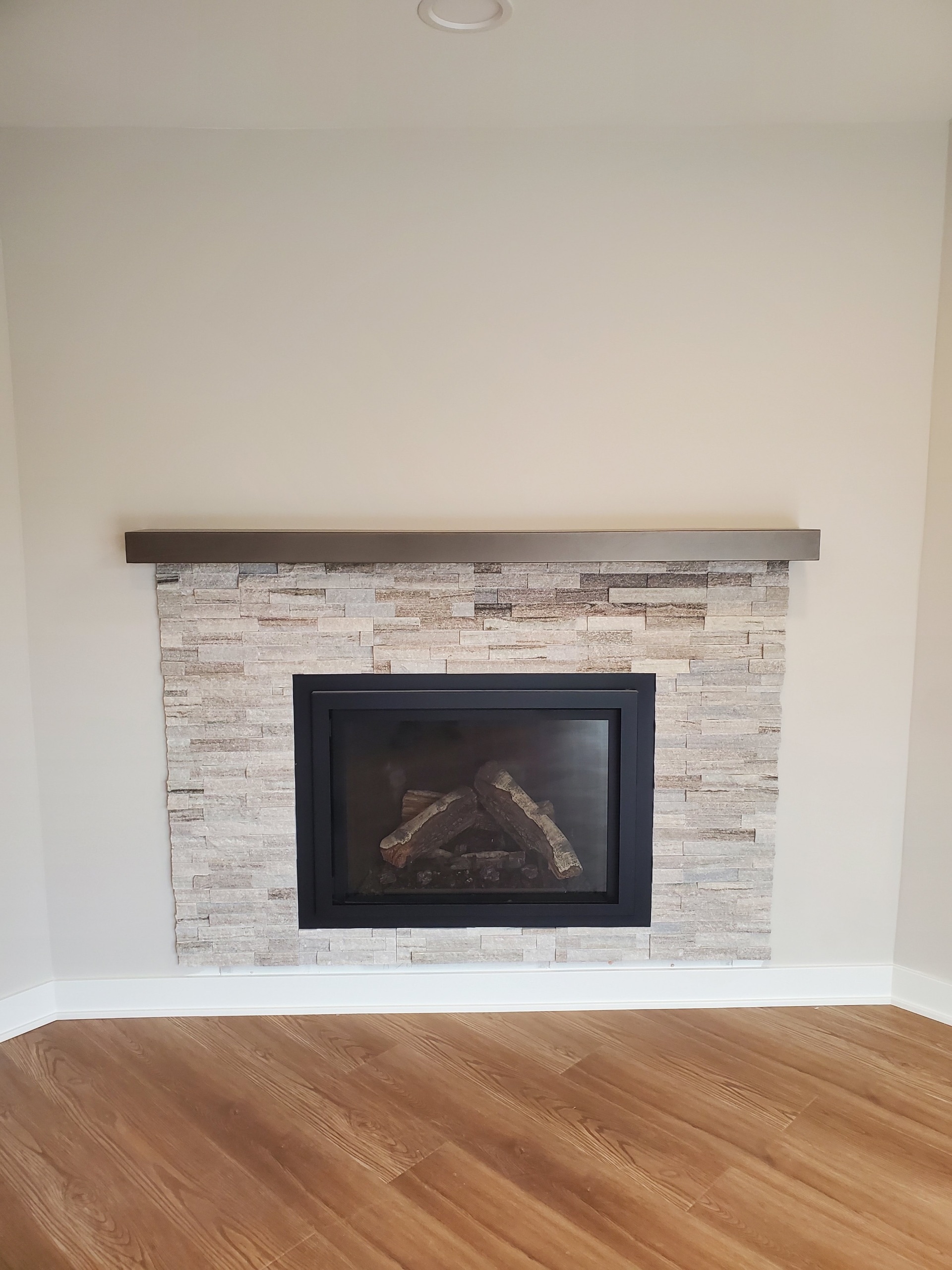 Modern linear electric fireplace with black frame and stacked stone surround in neutral tones, featuring a dark floating mantel, installed in a freshly painted living room by Chris Painting and Remodeling.