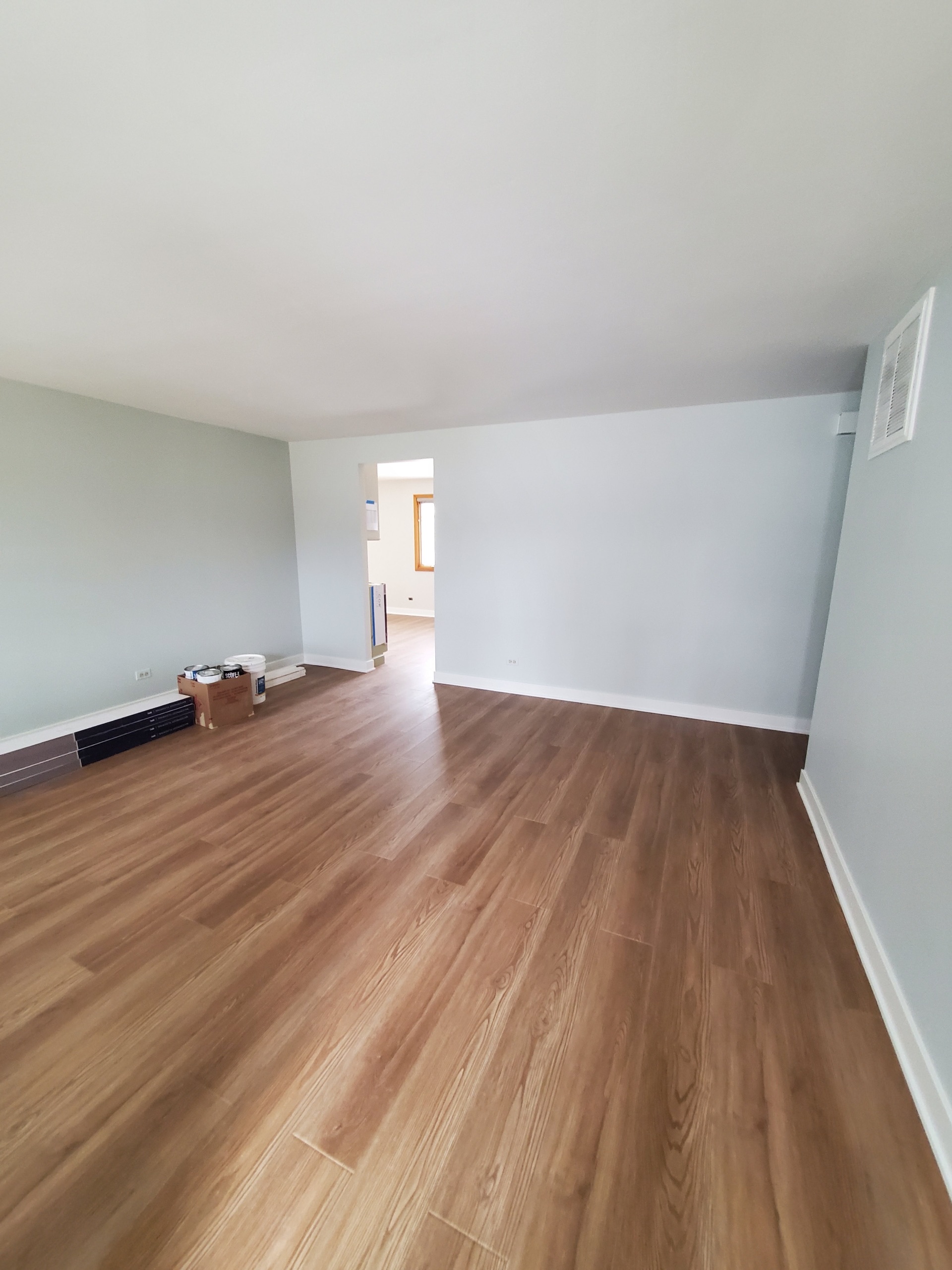 Spacious empty livingroom with fresh medium-tone hardwood-style flooring, light blue-gray walls, recessed lighting, baseboard heater, and doorway to adjacent space during remodeling by Chris Painting and Remodeling; paint cans and materials in corner.
