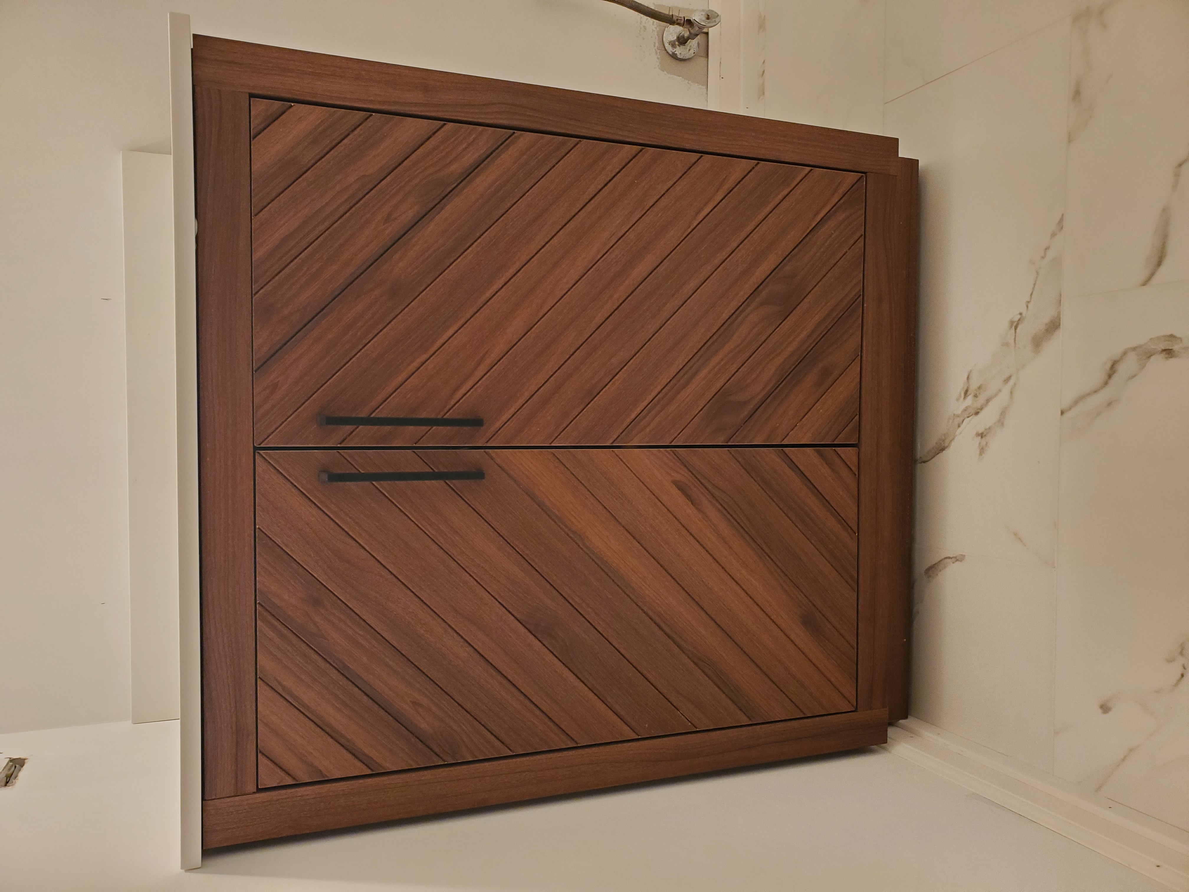 Close-up of walnut chevron-pattern bathroom vanity cabinet with matte black bar pulls and white quartz countertop on marble-look porcelain floor by Chris Painting and Remodeling.