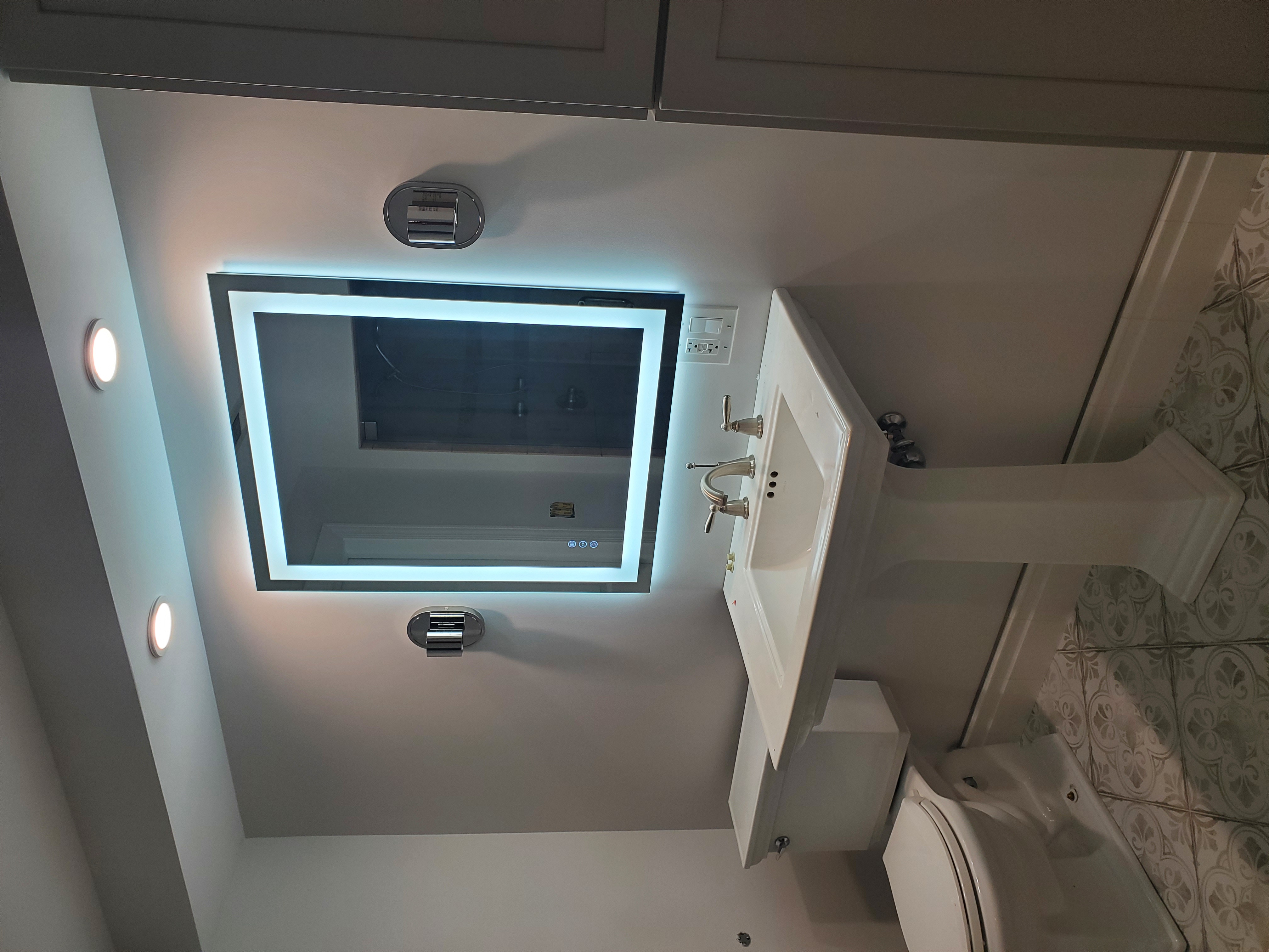 Remodeled bathroom with LED-backlit vanity mirror, white pedestal sink, brushed nickel fixtures, chrome wall sconces, and decorative patterned floor tile by Chris Painting and Remodeling.