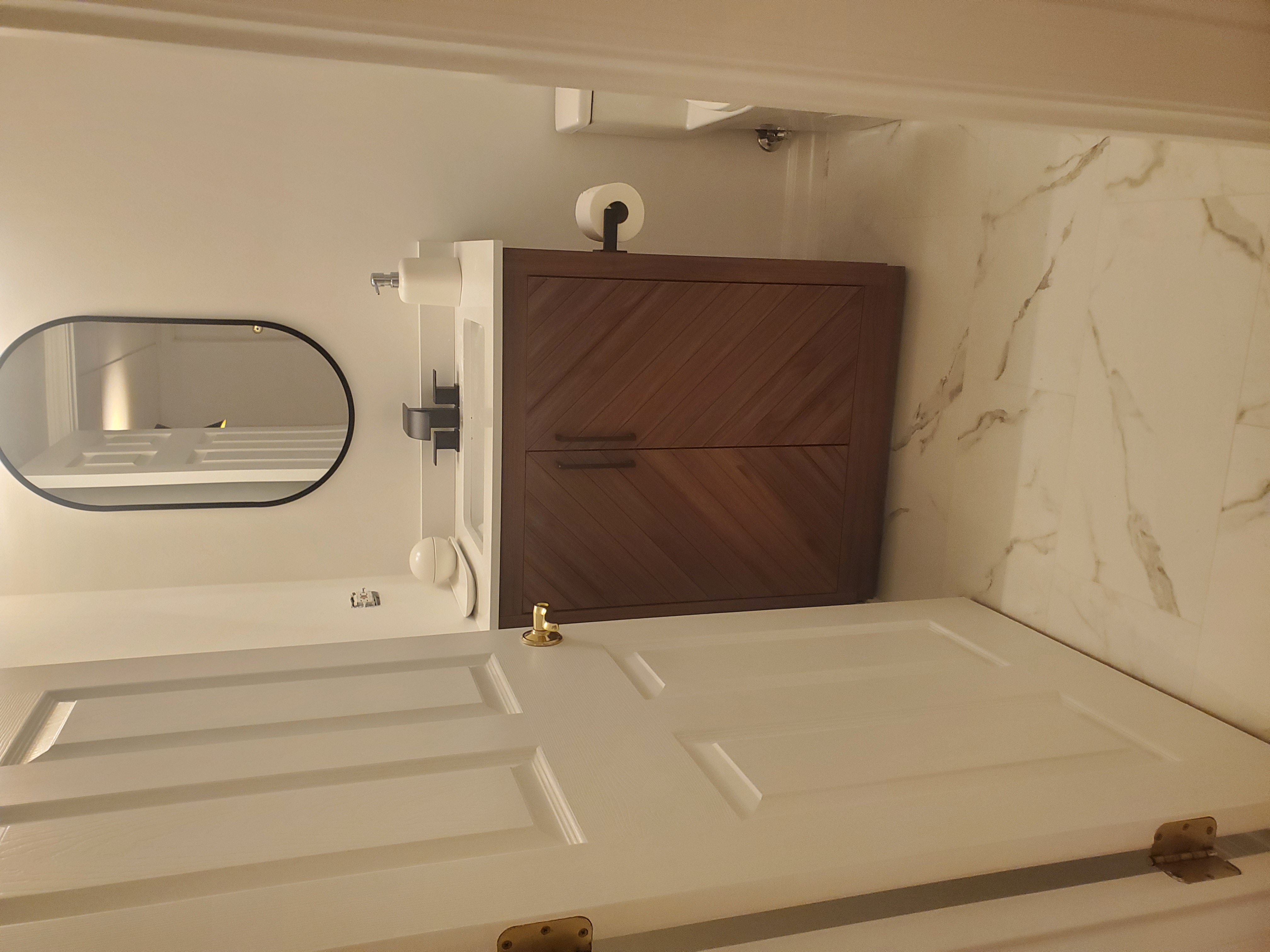 Remodeled half bath with walnut chevron-pattern vanity, white quartz top, matte black faucet, black-framed oval mirror, and marble-look porcelain floor tile by Chris Painting and Remodeling.