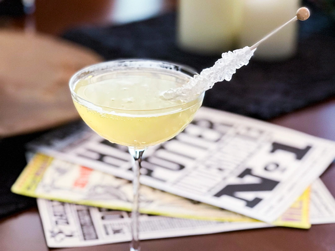 Yellow cocktail in a coupe glass garnished with a stick of rock candy, placed on a table with newspapers underneath.