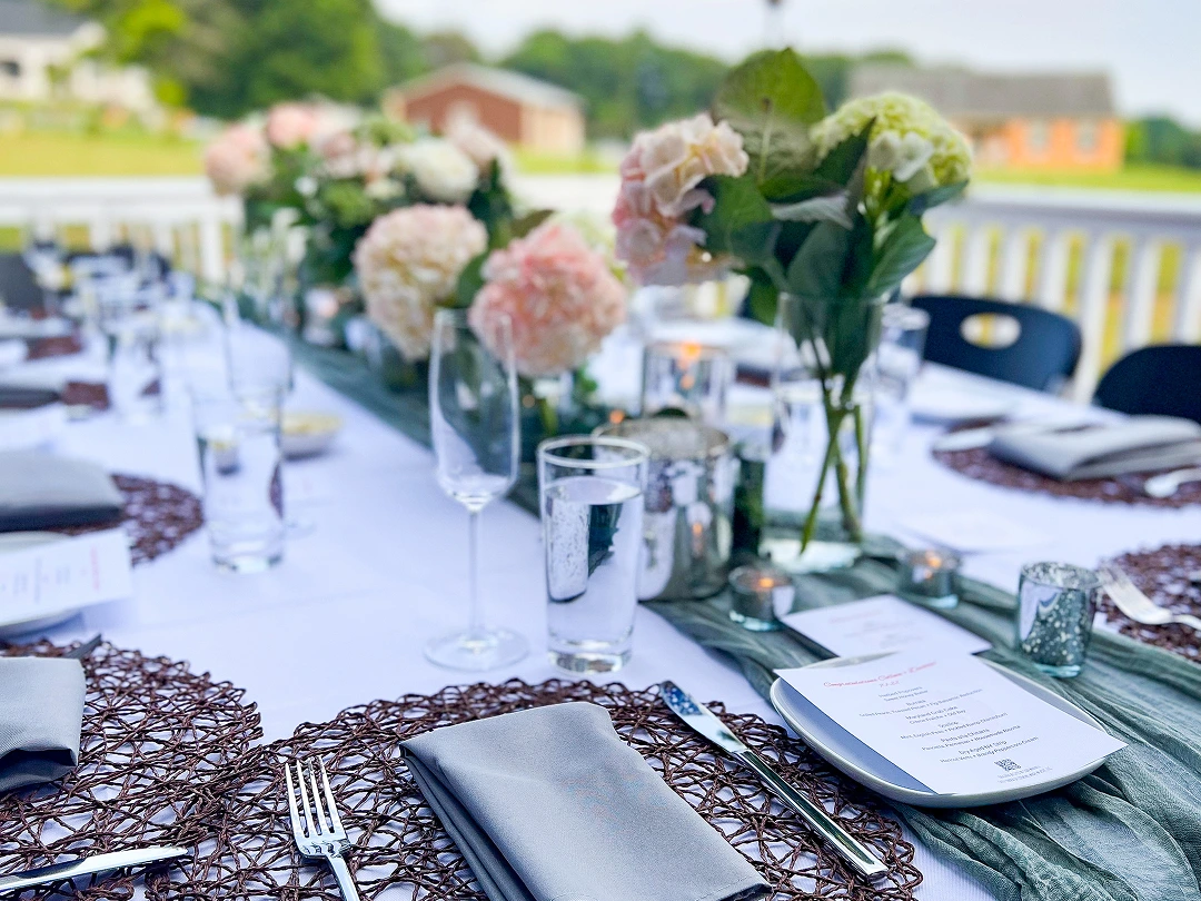 Elegant table setting with white tablecloth, woven placemats, gray napkins, crystal glasses, and floral centerpieces featuring pink and white flowers.