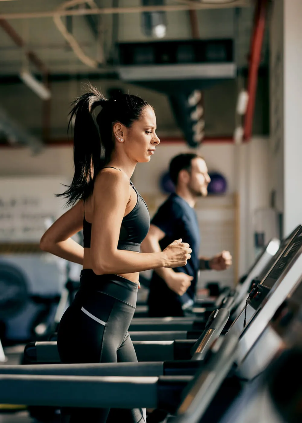 Woman running on treadmill in modern gym, focused on fitness and exercise, with another person jogging nearby, illustrating active lifestyle and gym environment.