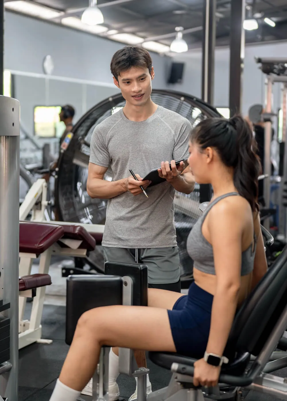 Personal trainer assisting a female client in a gym, discussing workout techniques while she uses a leg press machine, emphasizing fitness training and professional development in the fitness industry.