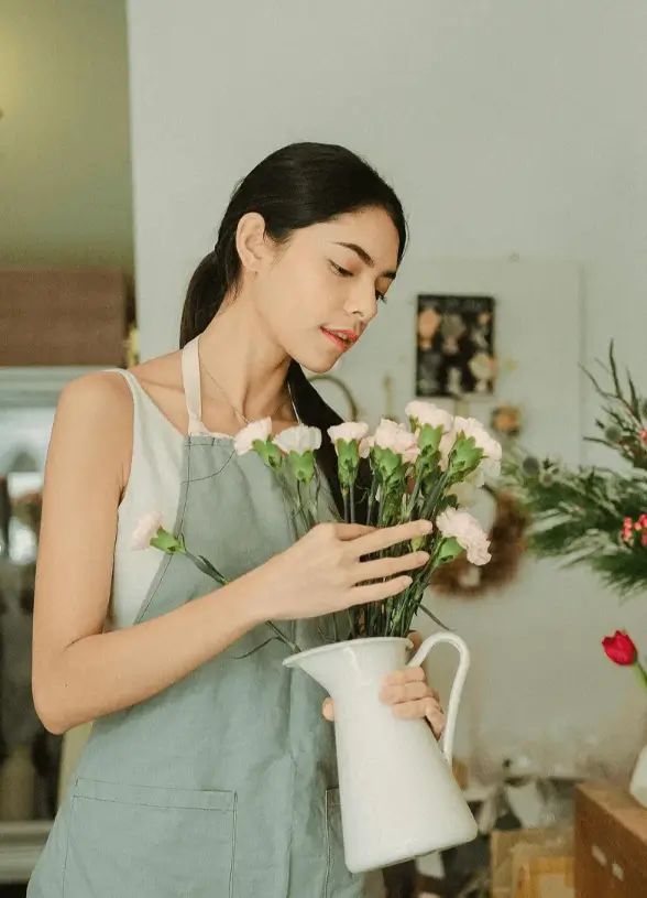 Woman in apron arranging pink flowers in a white vase, representing creativity and business growth in a floral shop setting.