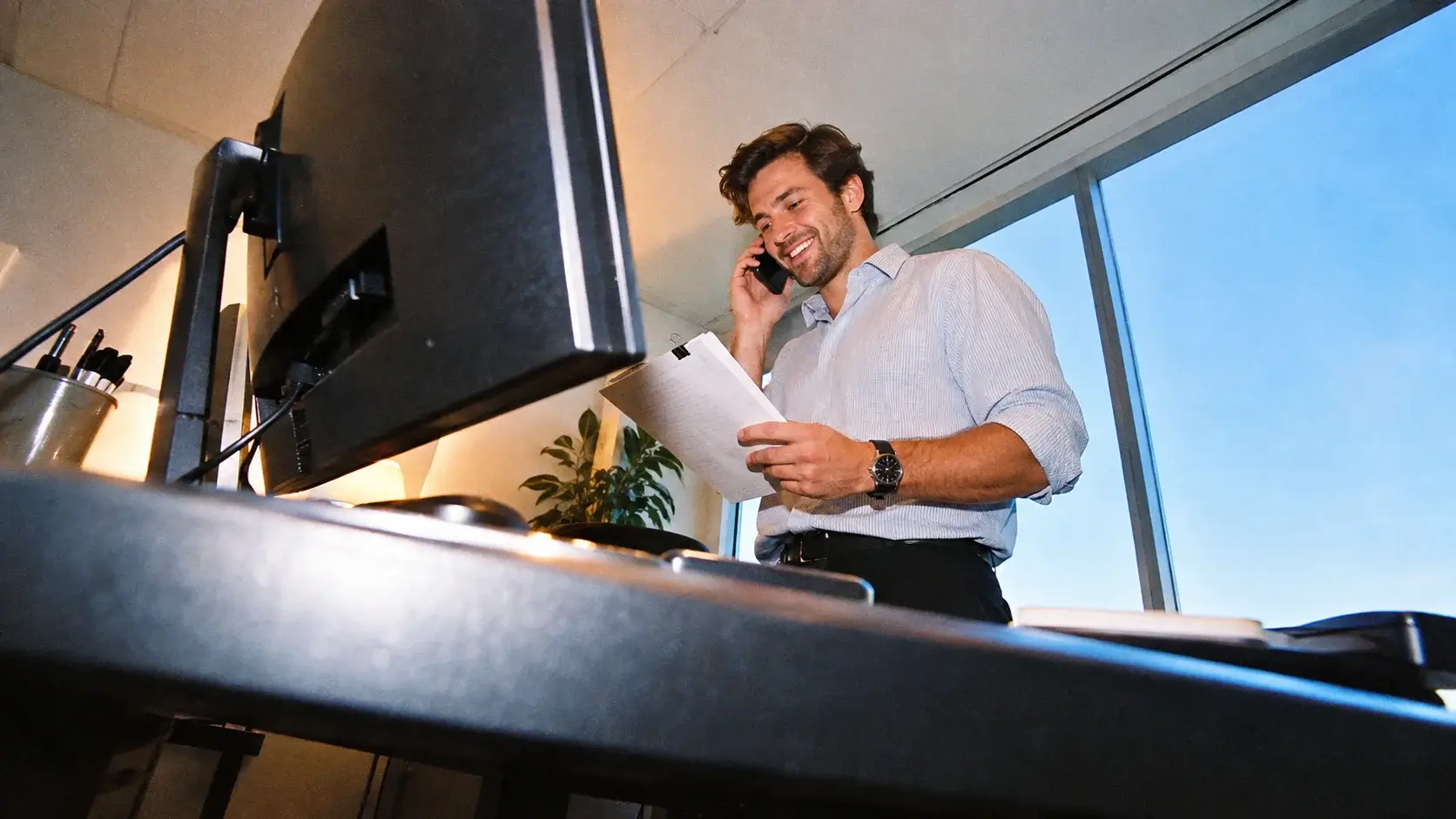 Recruiter smiling on a phone call while holding paperwork beside a window in a high-rise office