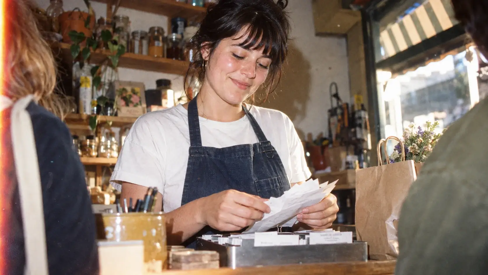 Café owner in a denim apron smiling while sorting receipts at the counter