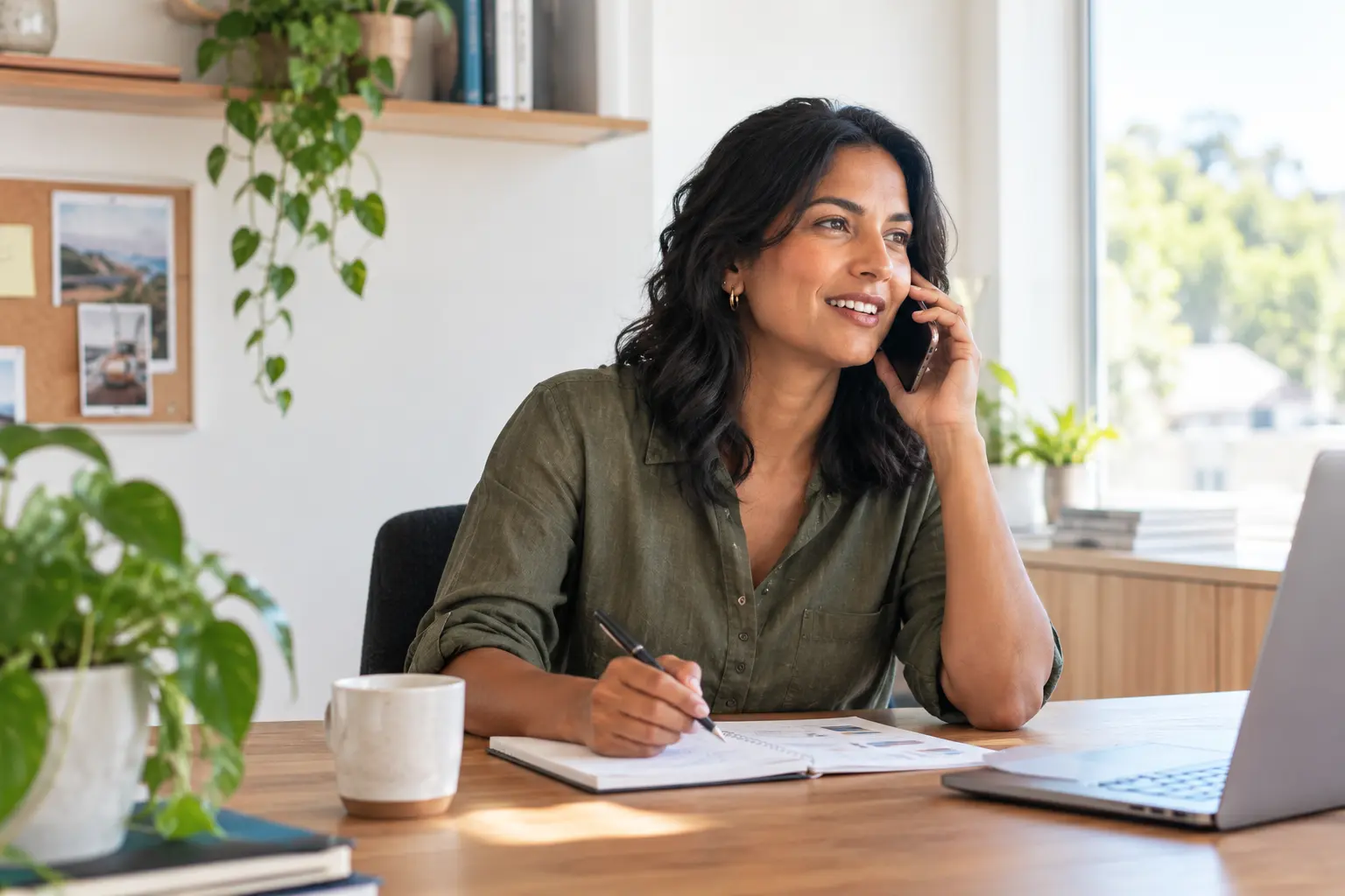 A casually dressed finance broker speaking on the phone at a desk in a bright, relaxed home office, with a notebook and laptop nearby.