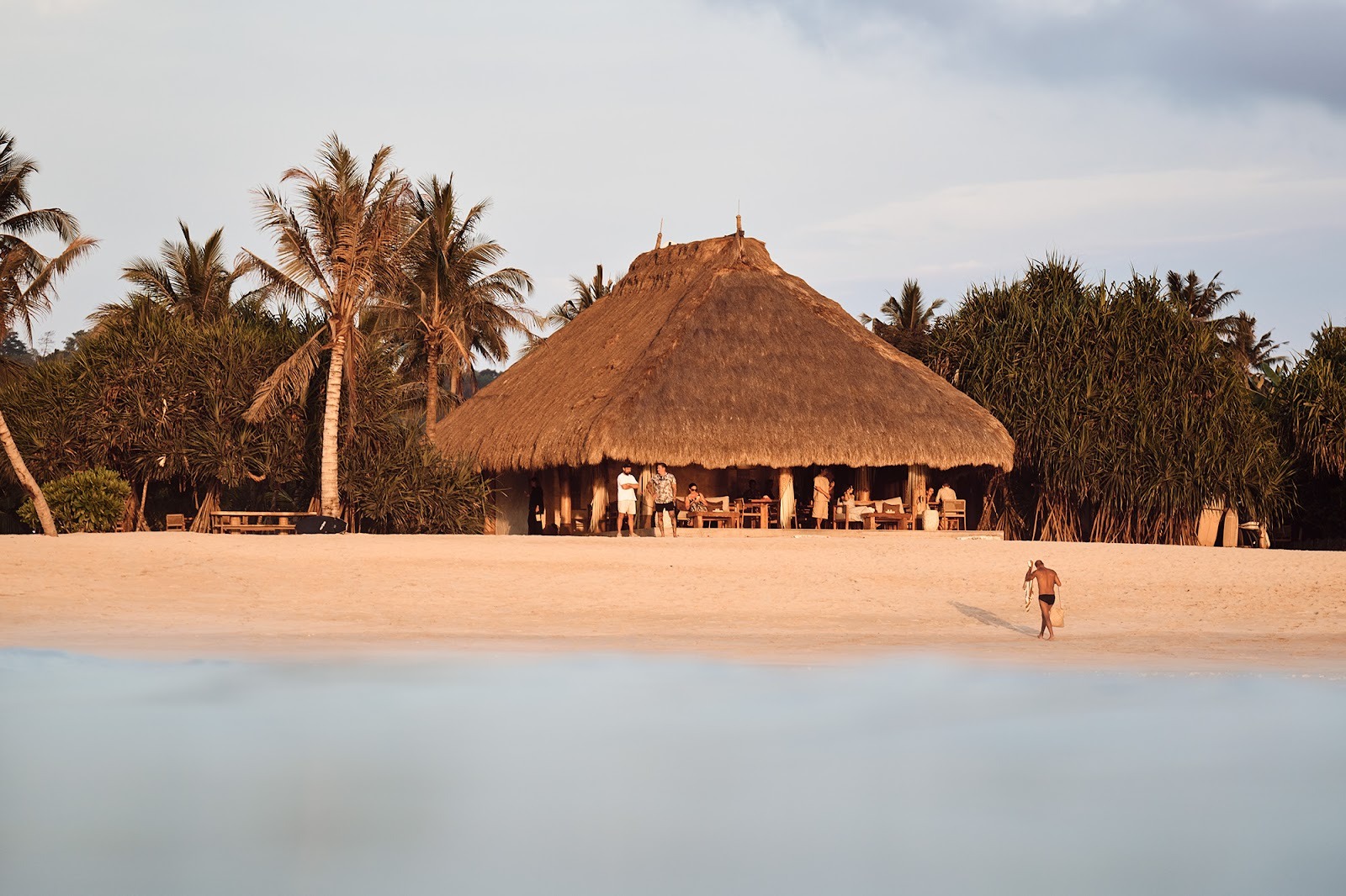 Beachfront bamboo hut restaurant in Sumba with people relaxing by the sea during sunset.