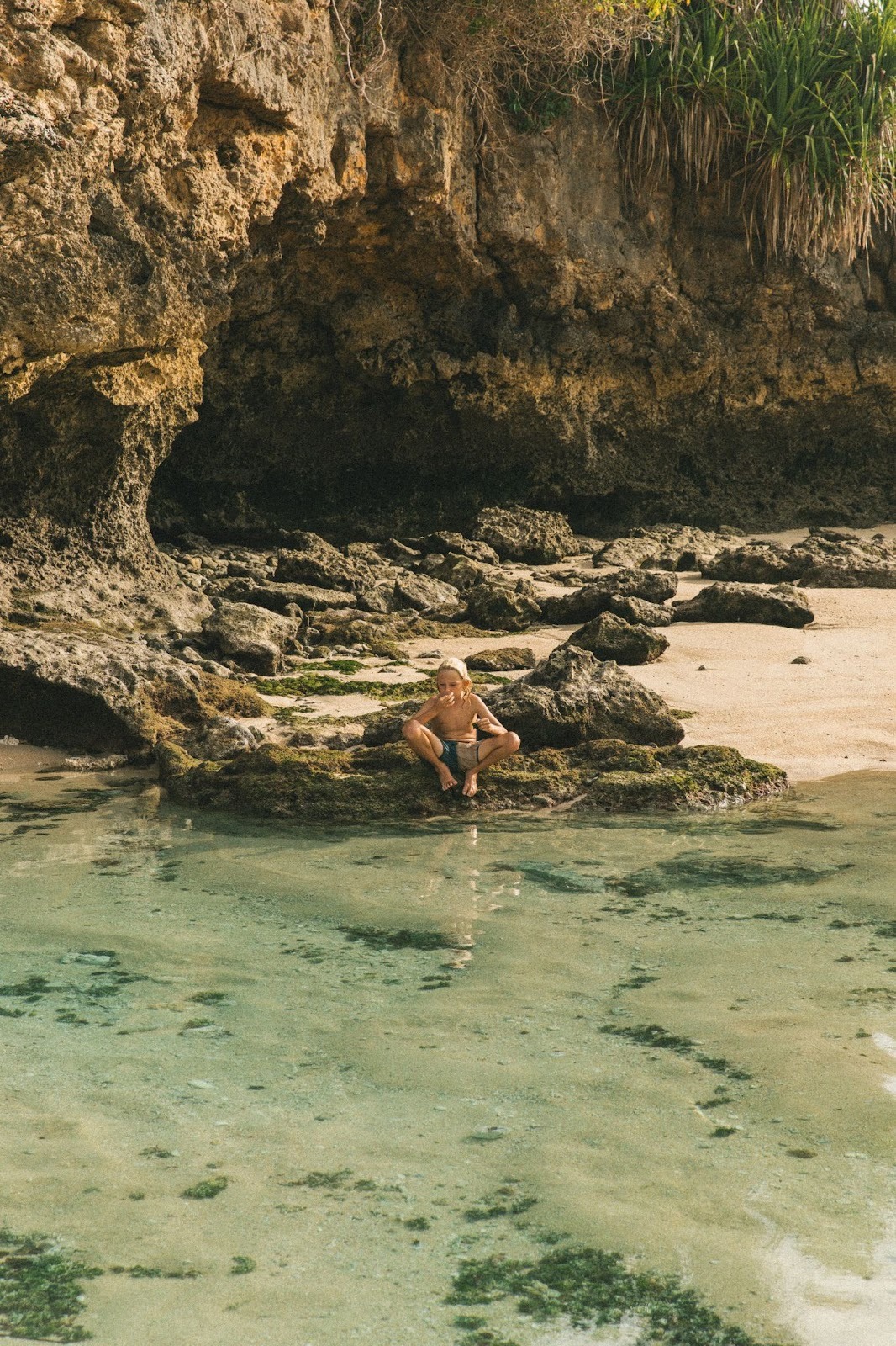 A boy sitting between a rock at clear and calm natural beach pool