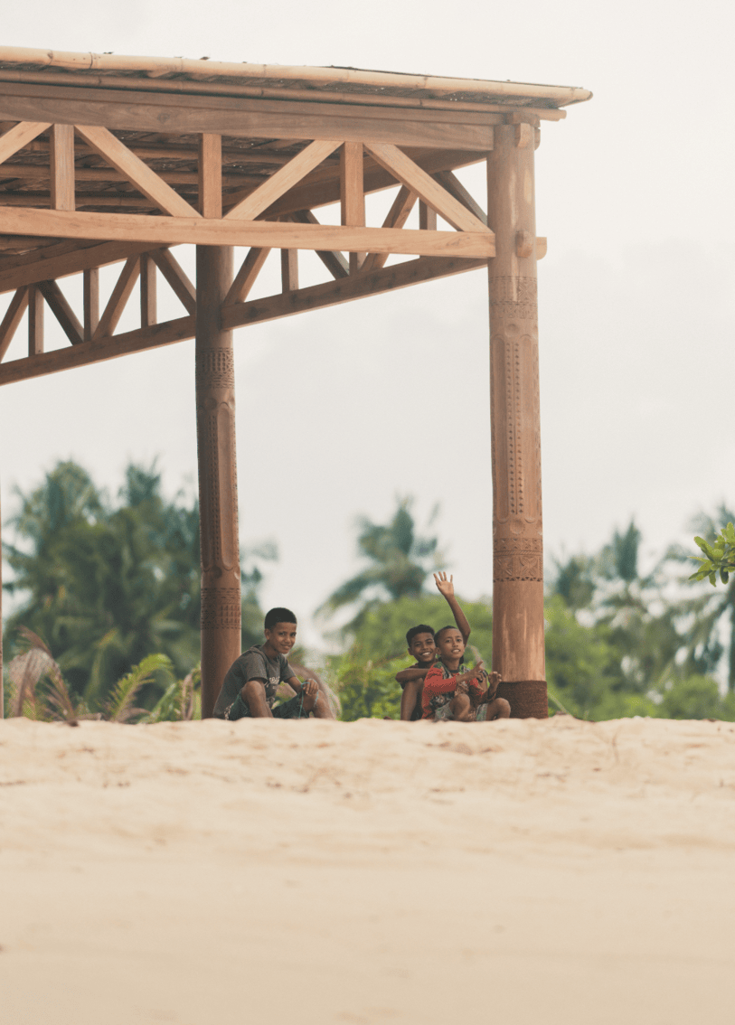 Children in West Sumba sitting under traditional wooden structure near the beach reflecting local community life and sustainable tourism impact