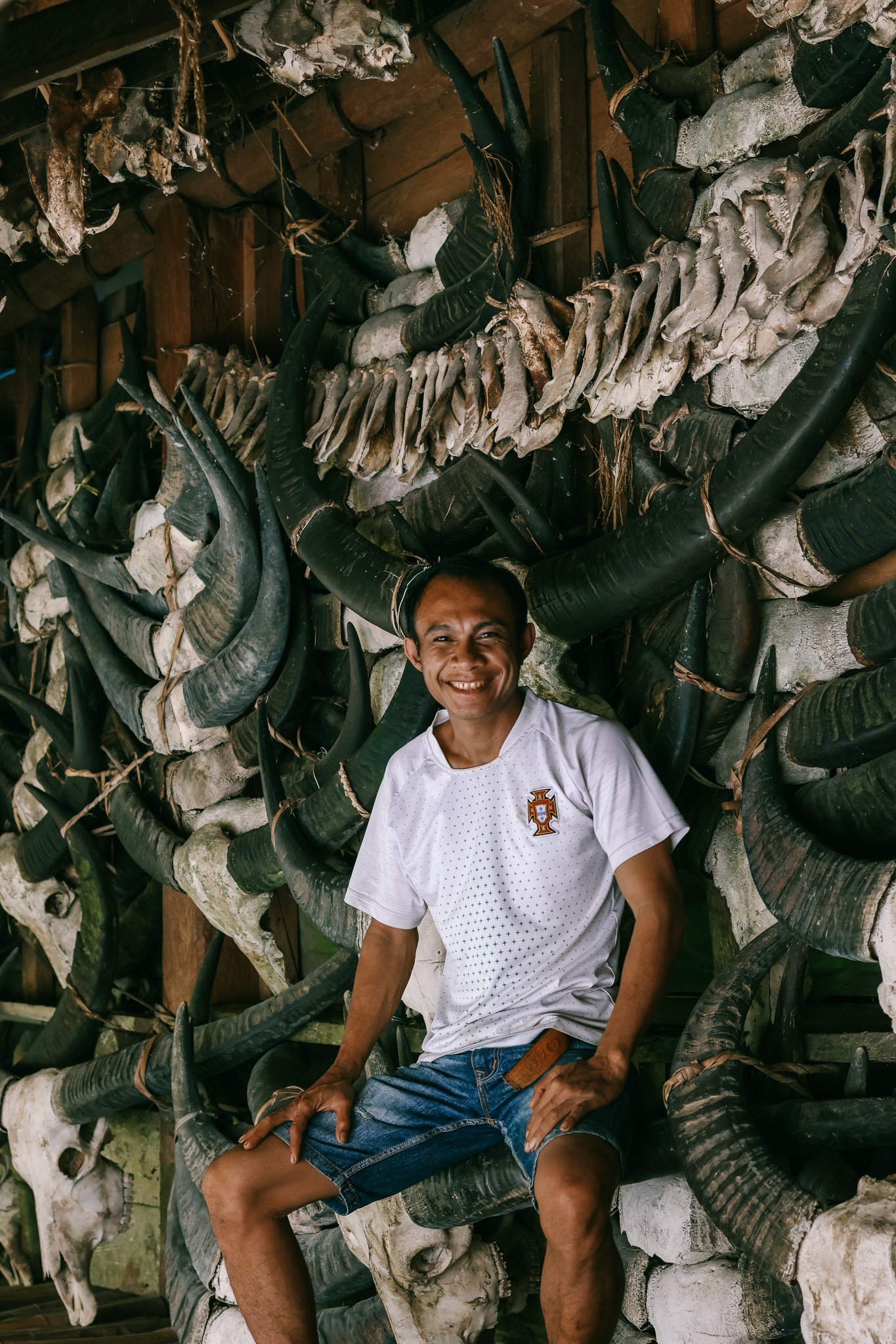 Local Sumbanese man sitting in front of a traditional village house wall covered in water buffalo skulls and horns — Marapu ancestral heritage West Sumba Indonesia