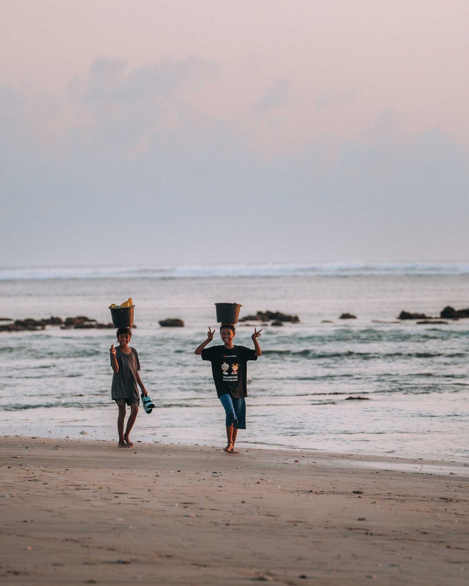 Two young Sumbanese women carrying water buckets on their heads along a beach in West Sumba Indonesia — water collection daily life near The Sanubari Sumba