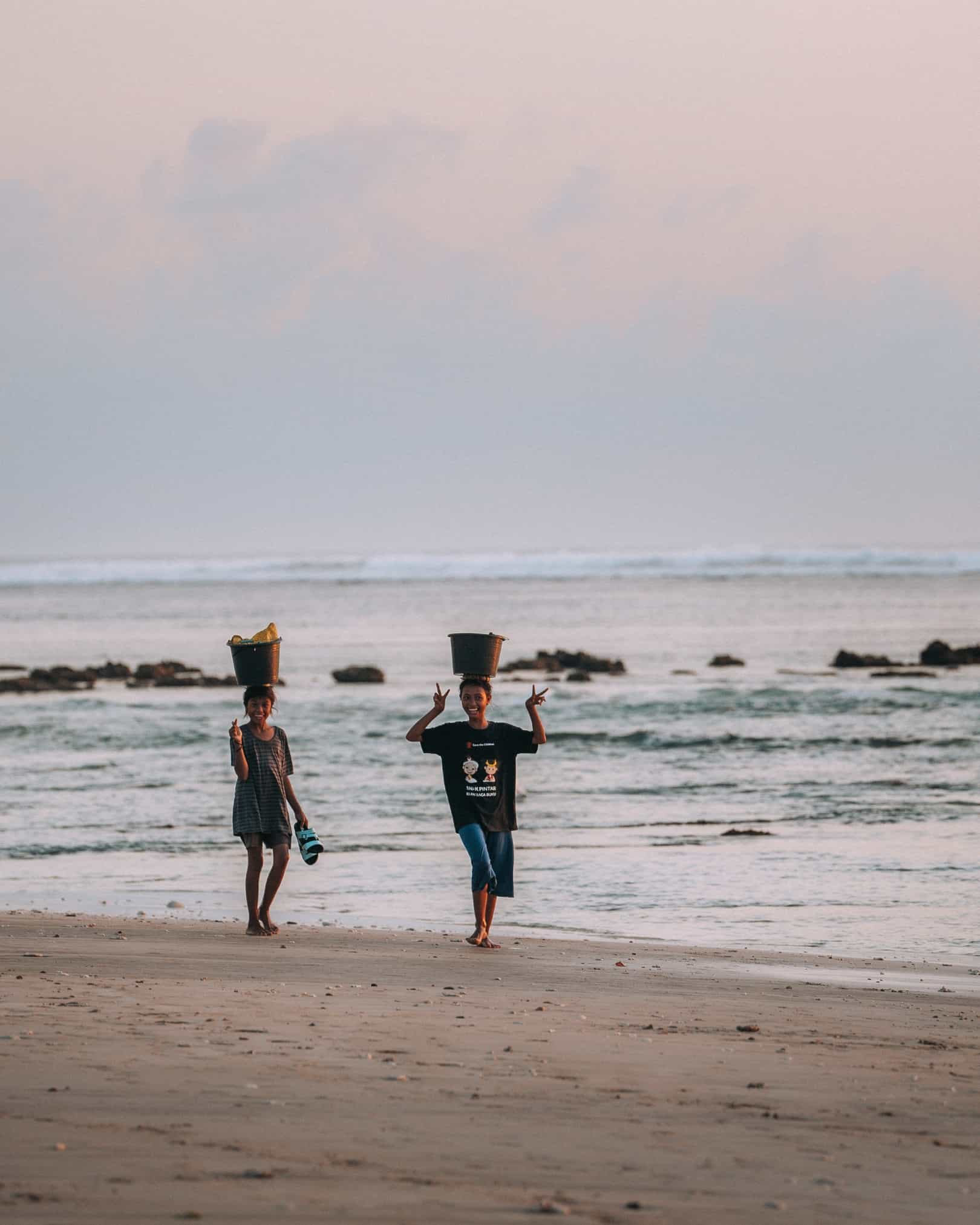 Two young Sumbanese women carrying water buckets on their heads along a beach in West Sumba Indonesia — water collection daily life near The Sanubari Sumba