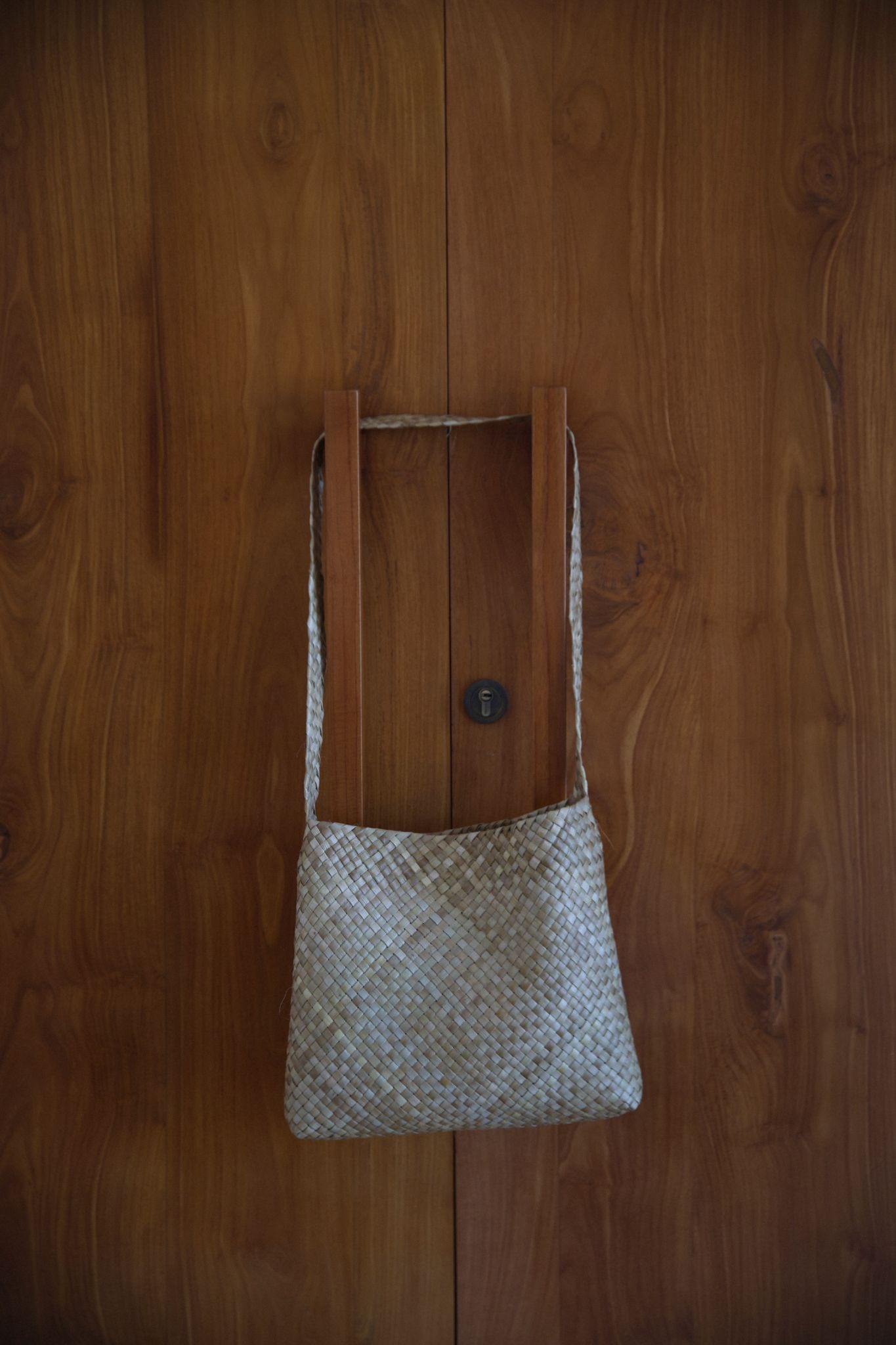 A biodegradable, hand-woven pandan leaf bag hanging on a wooden door, available as an authentic cultural souvenir at The Sanubari Sumba boutique.