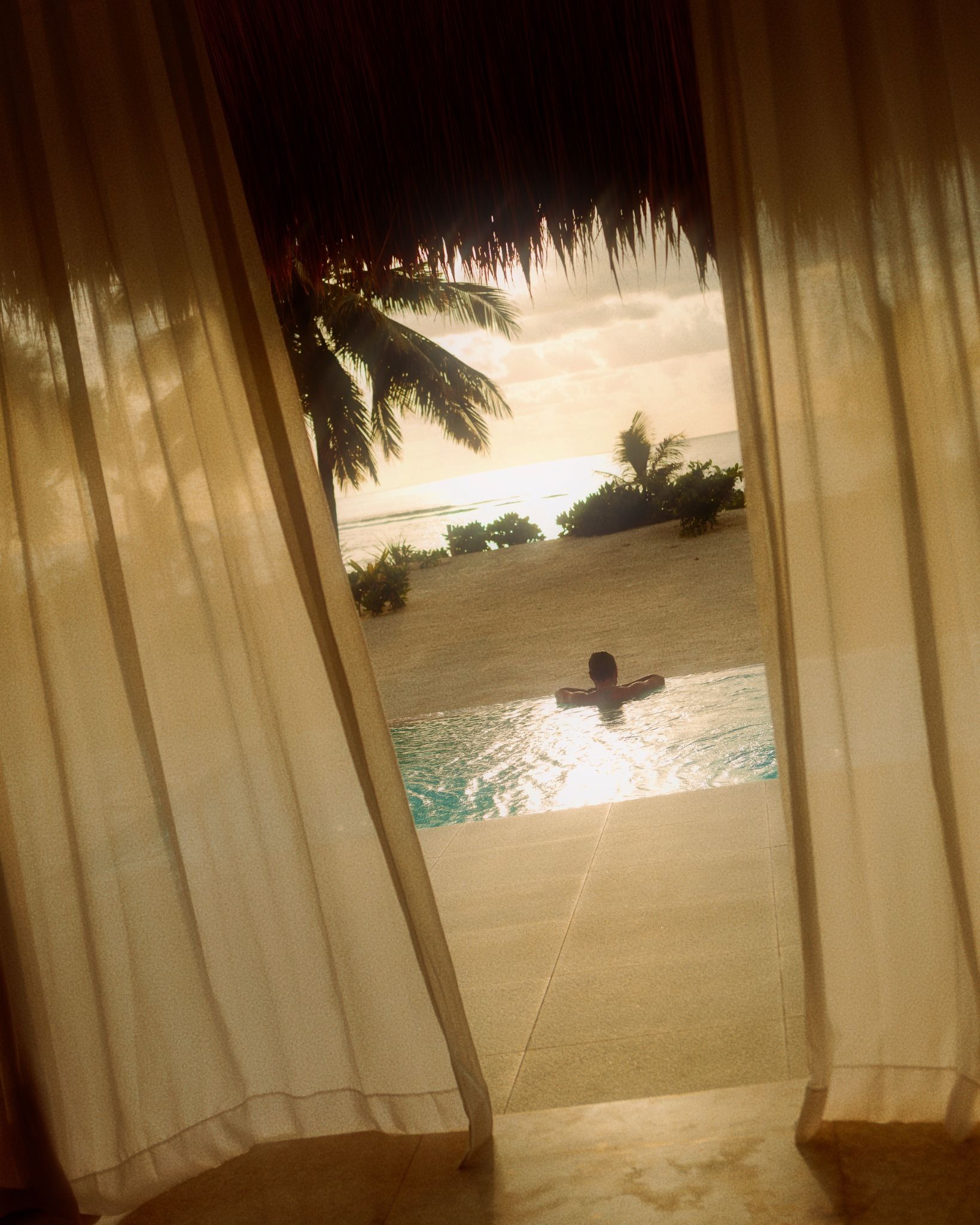 A guest relaxing in a private infinity pool overlooking the beach at sunset, framed by sheer curtains in a luxury beachfront villa at The Sanubari Sumba.