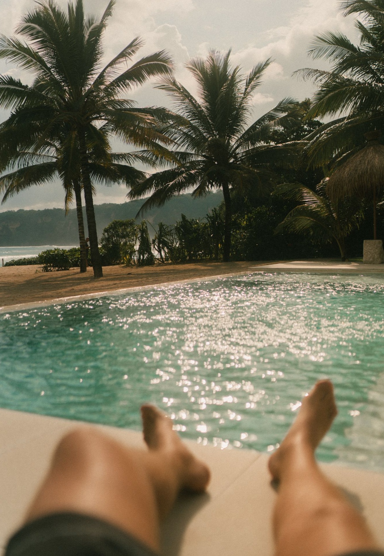 First-person view relaxing poolside under towering coconut palm trees with ocean views in Sumba.