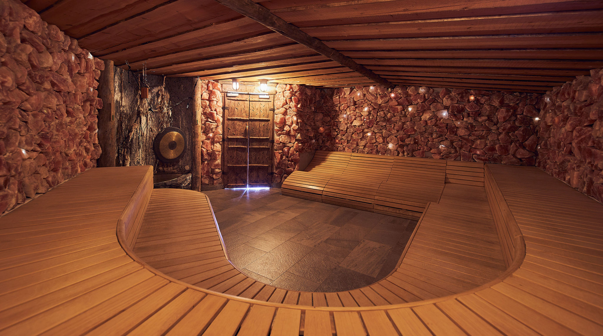 Un homme profite de l'eau d'un étang de baignade naturel tandis qu'une femme vêtue d'une serviette blanche est assise sur un rocher et sourit, avec un cadre naturel verdoyant en arrière-plan.