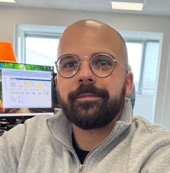 Man met een kaal hoofd, bril en baard draagt een lichtgrijze trui en kijkt naar de camera in een kantooromgeving met computerscherm op de achtergrond.