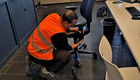 Man in oranje veiligheidsvest knielt en repareert een bureaustoel in een kantoorruimte.