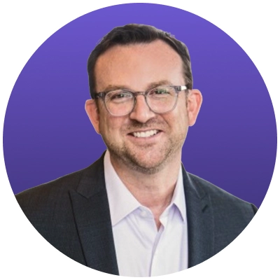 Smiling man with glasses, short hair, and beard wearing a dark blazer and white shirt on purple background.