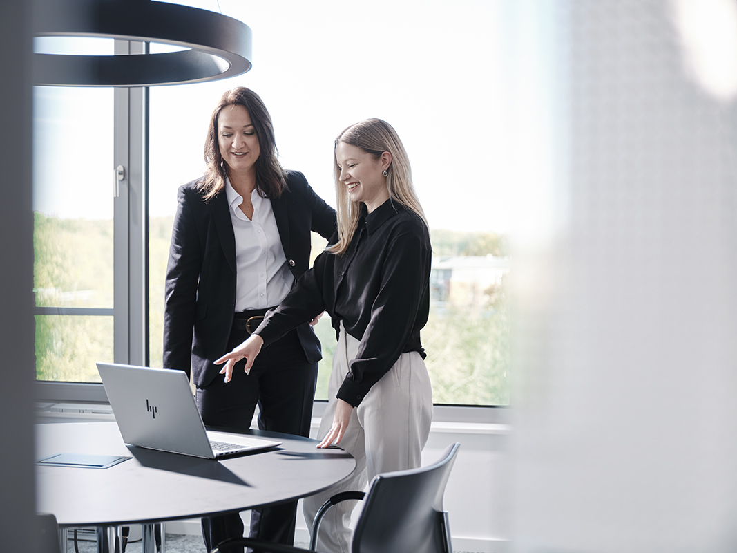 Zwei Frauen stehen an einem runden Tisch vor einem geöffneten Laptop in einem hellen Büro mit großen Fenstern.