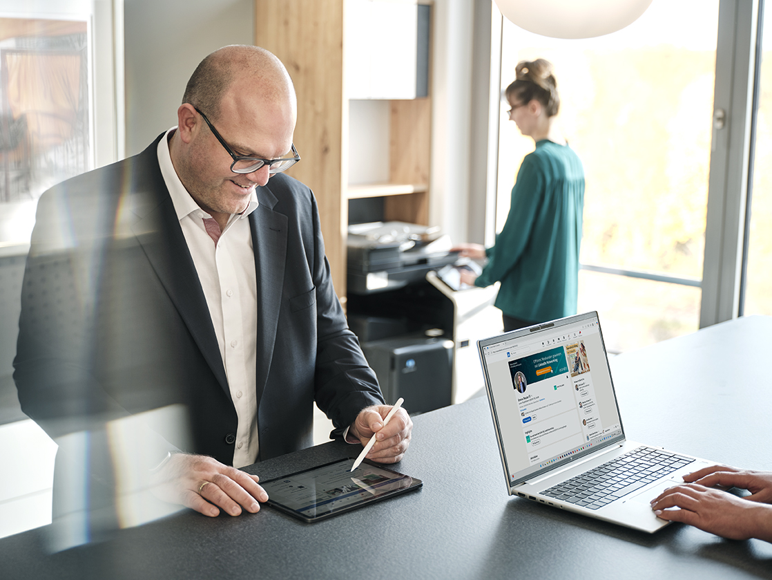 Ein Mann in Anzug steht an einem Tresen und schreibt mit einem Stift auf einem Tablet, während eine Frau im Hintergrund an einem Drucker arbeitet und eine weitere Person an einem Laptop tippt.