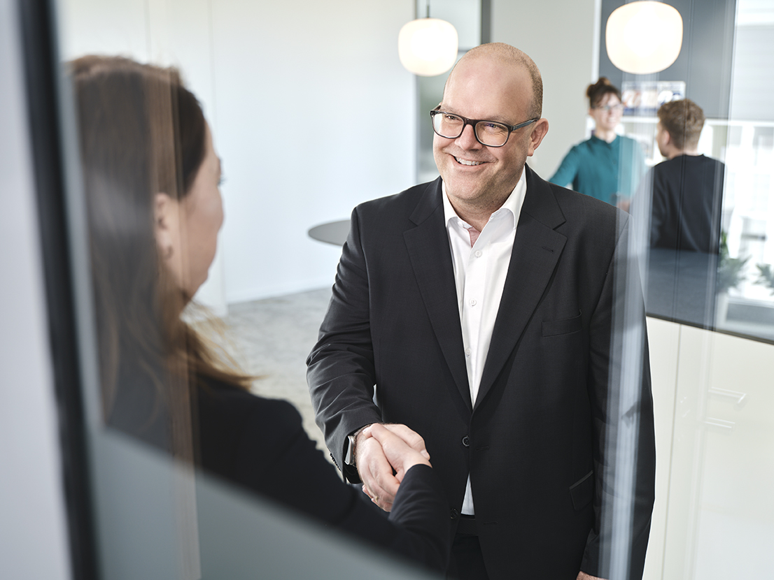 Zwei Personen geben sich die Hand in einem modernen Büro mit weiteren Personen im Hintergrund