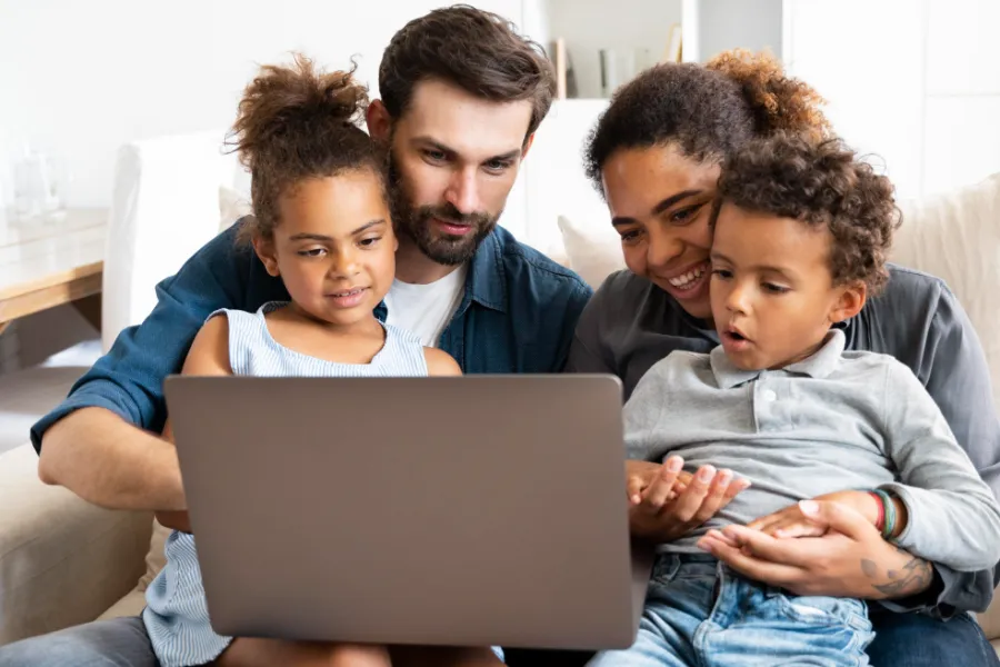 A happy family of four sitting together on a couch, engaging with content on a laptop screen in a cozy living room.