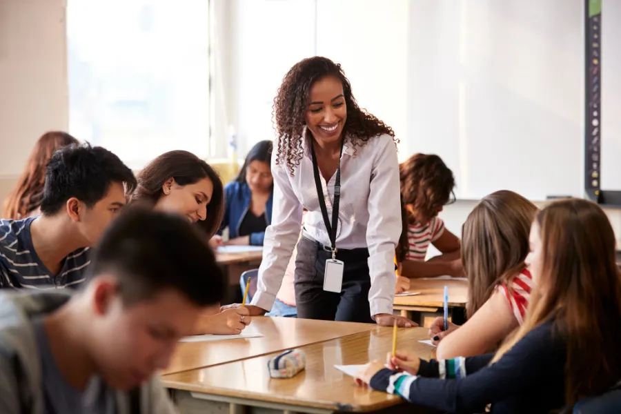 Smiling teacher engaging with students working at their desks in a bright, collaborative classroom setting.
