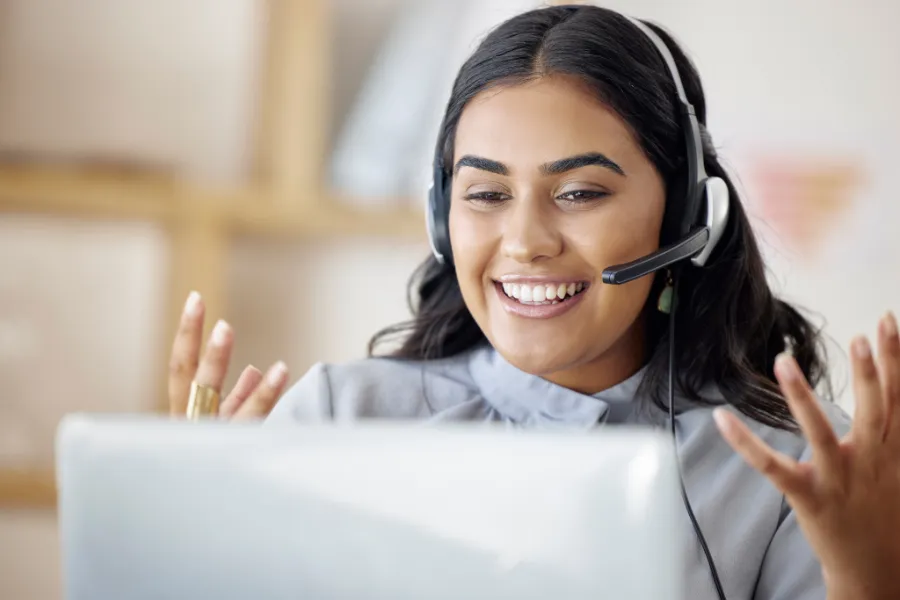 Smiling woman wearing a headset during a virtual meeting, gesturing with her hands in a bright office space.