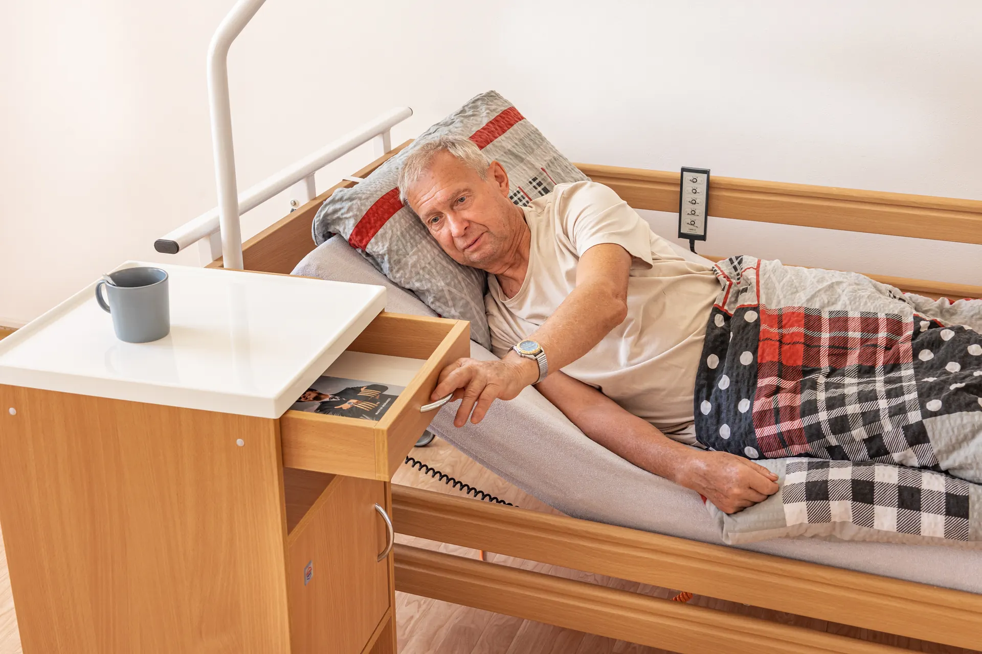 Senior na polohovací posteli si bere věc ze zásuvky nočního stolku