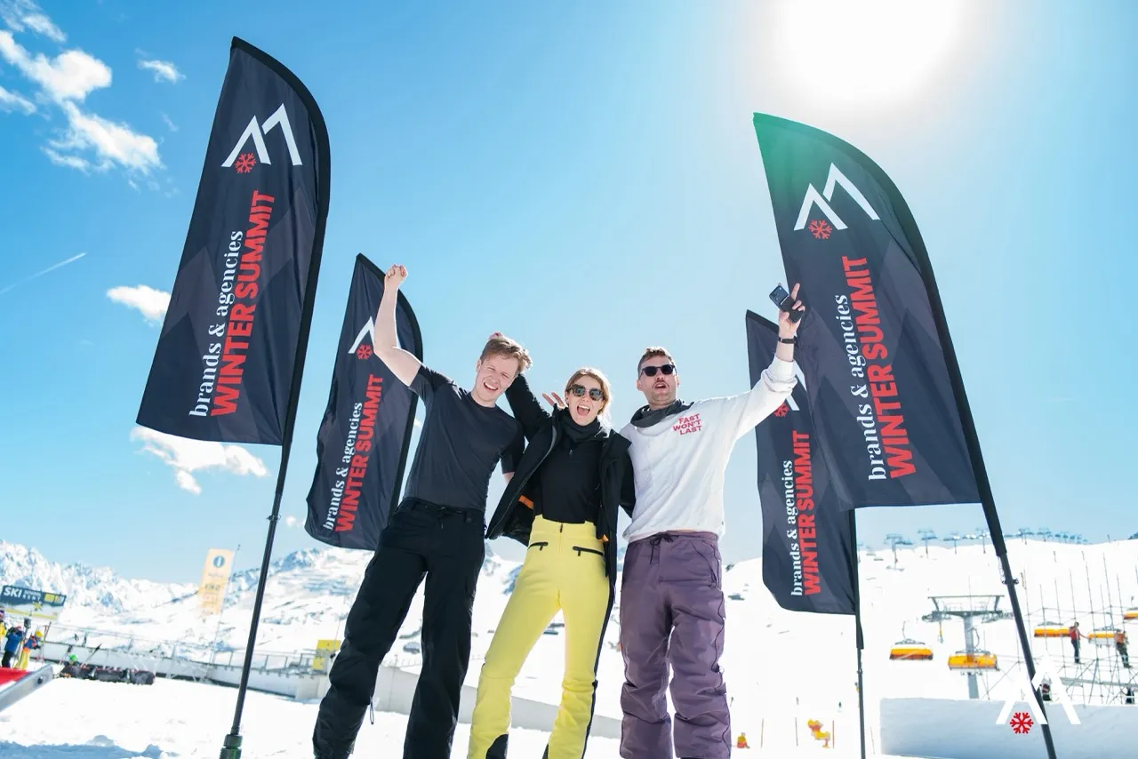 Drei Teilnehmende jubeln zwischen den WINTER SUMMIT Flags im sonnigen Skigebiet