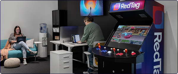 A woman sitting on the couch with a laptop and another standing and working at her desk next to an arcade machine in an office