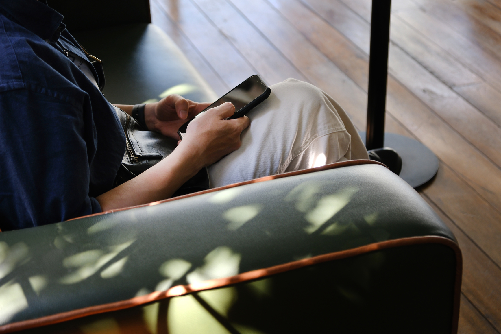 Young man wears blue tshirt, sitting on green leather sofa, checking his phone under the shadow of sunlight.
