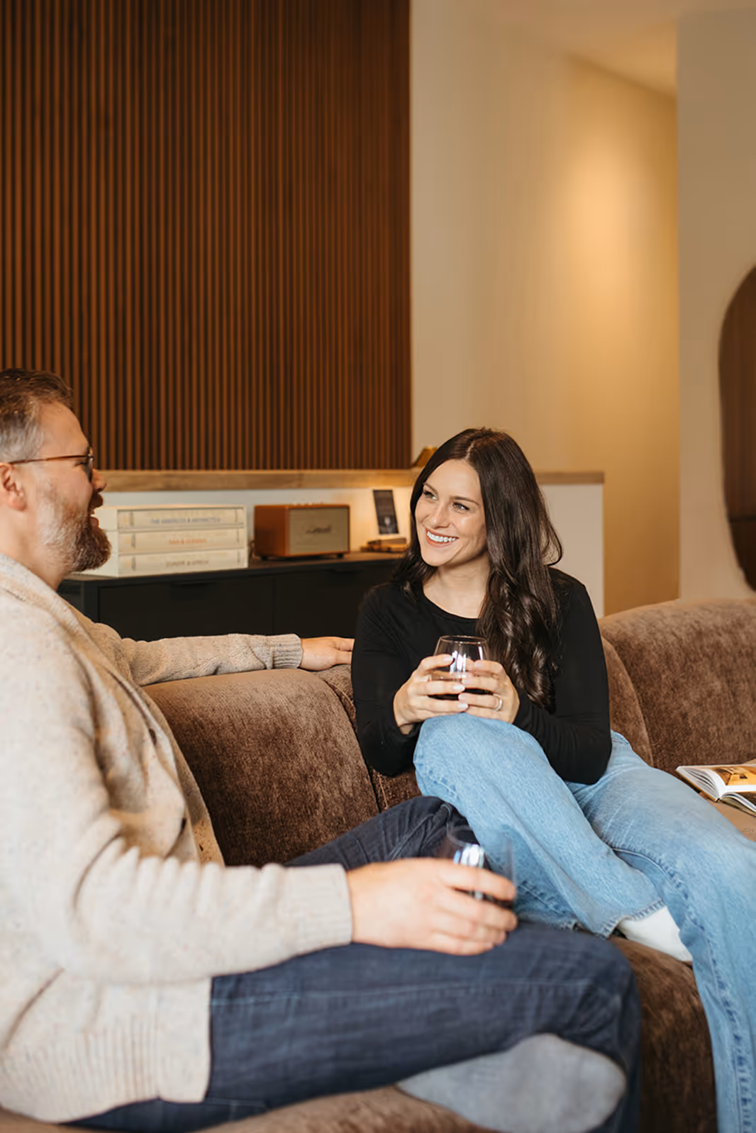 Man and woman sitting on a brown couch, smiling and holding glasses of red wine in a cozy living room.