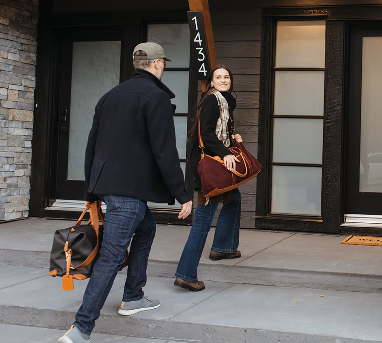 A man and woman carrying travel bags walking up steps to a house.
