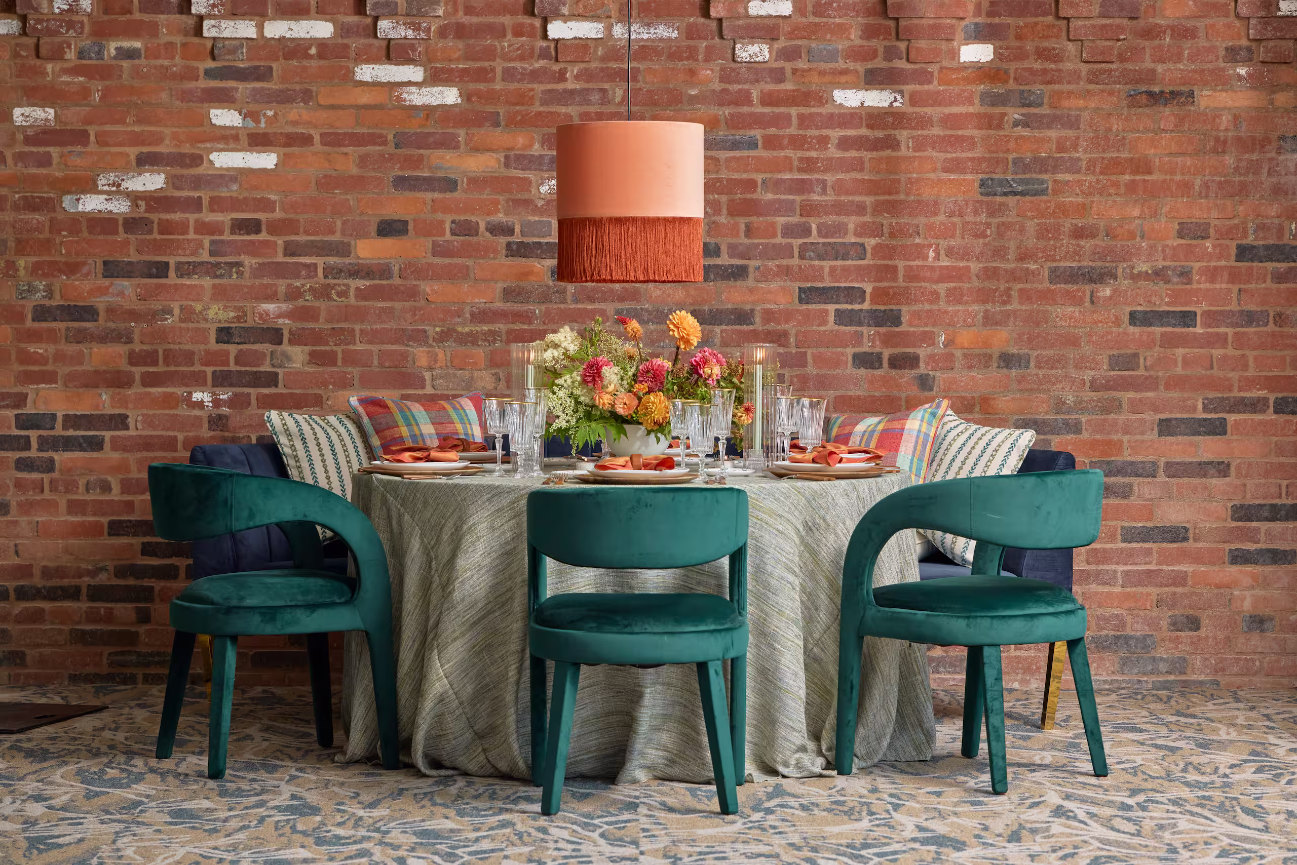 A contemporary bohemian table setting featuring green velvet chairs, red and orange flowers, a couch and a hanging orange lamp against an exposed brick wall. Photo by Adam Milliron