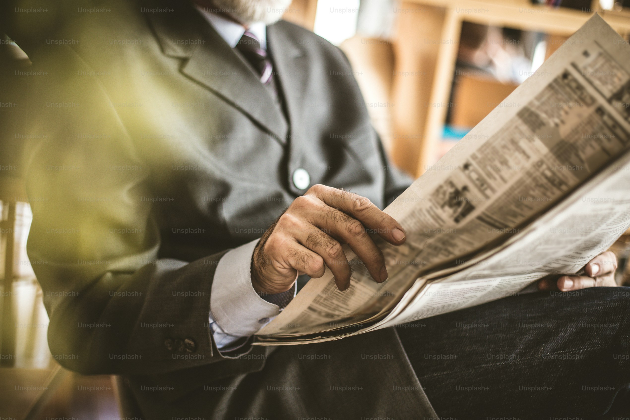 A man in a grey suit sitting reading a newspaper.