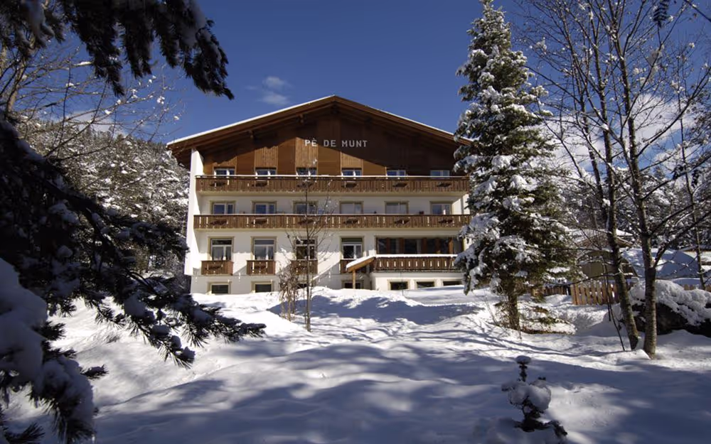Mehrstöckiges Holzhaus mit Balkon im Schnee, umgeben von schneebedeckten Bäumen unter blauem Himmel.