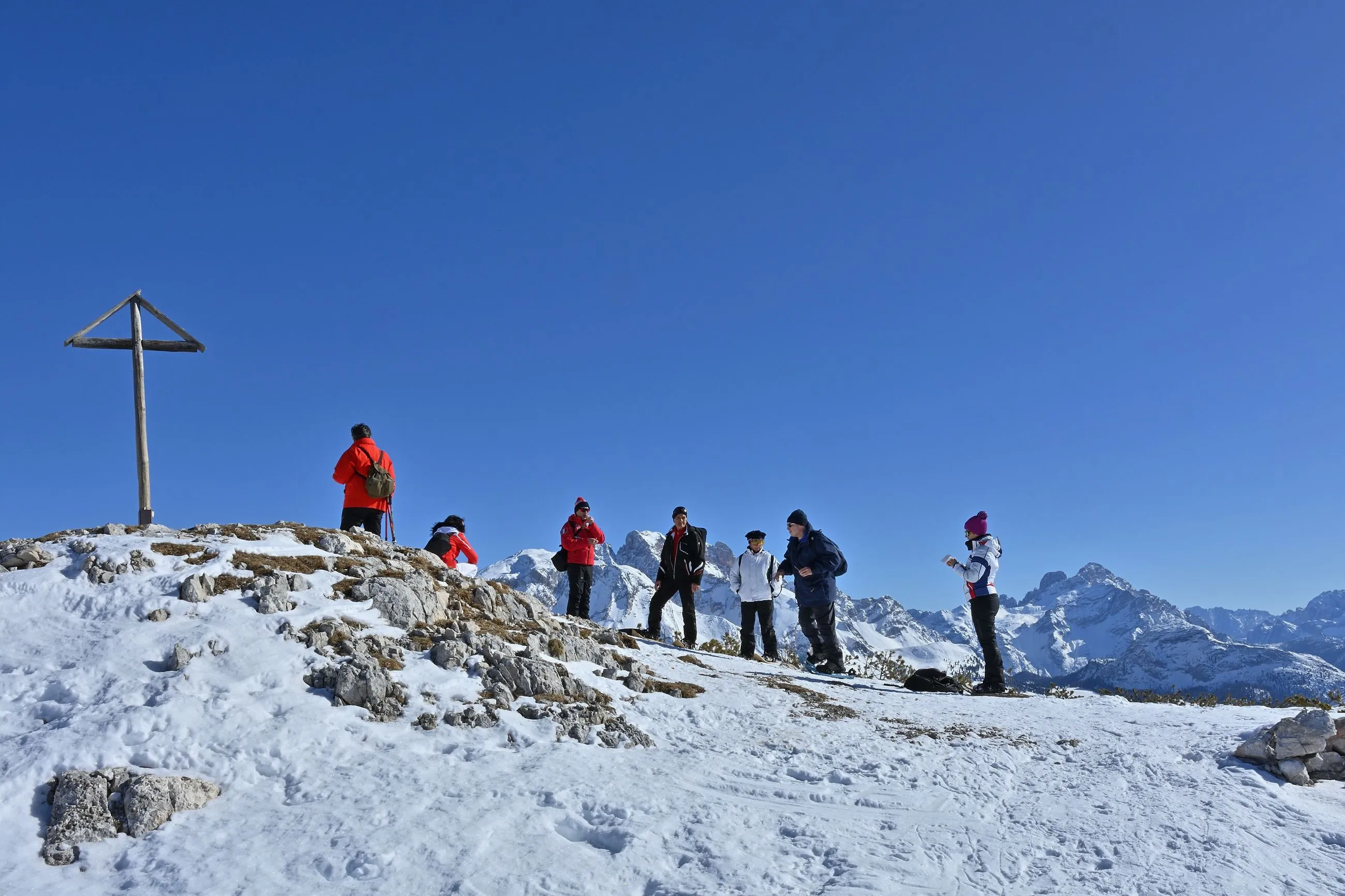 Gruppe von Wanderern in Winterkleidung auf einem schneebedeckten Gipfel mit hölzernem Gipfelkreuz und Bergpanorama im Hintergrund.